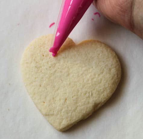 Gluten-free sugar cookie hearts being frosted with pink frosting.